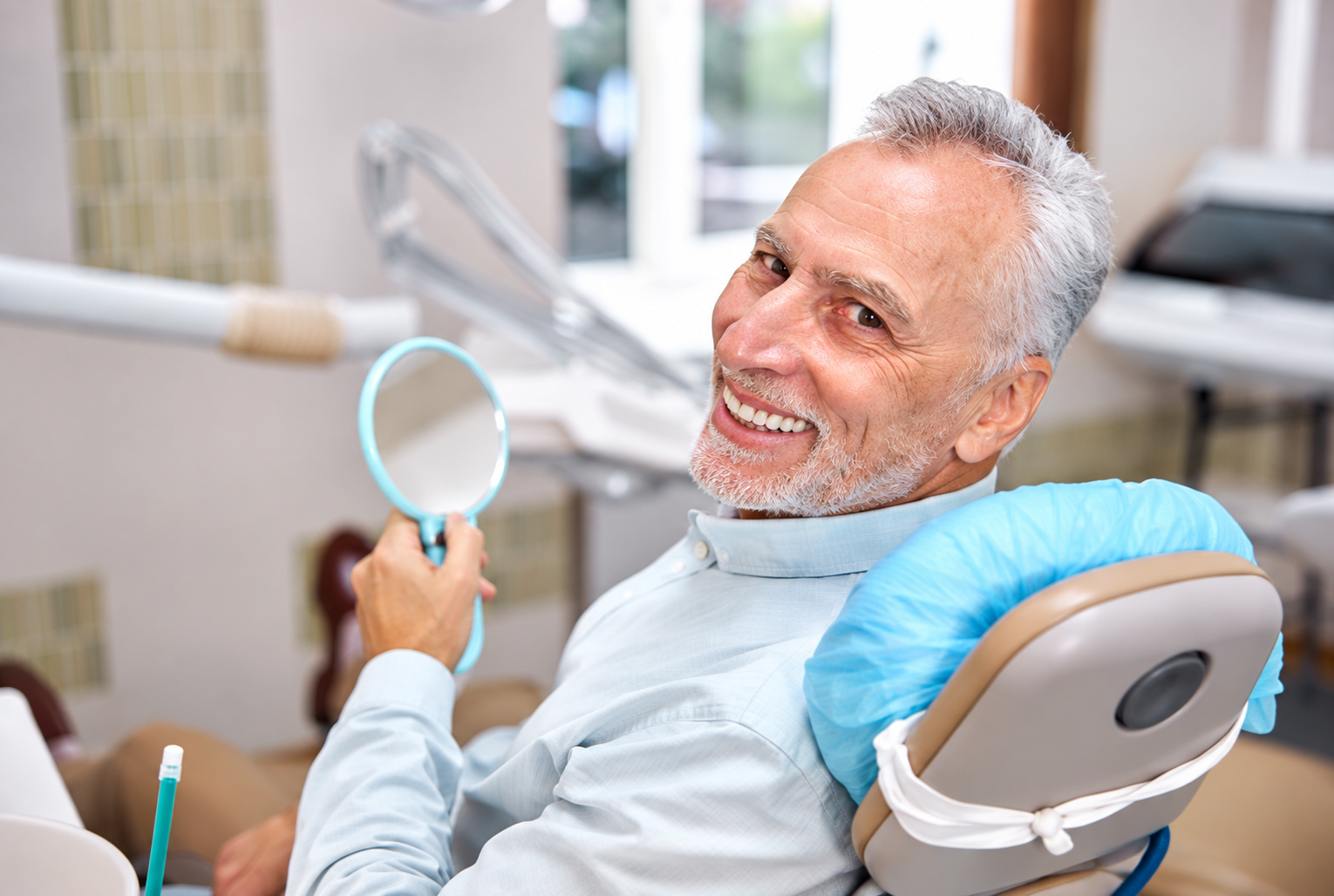 Smiling senior man with overdentures holding a dental mirror in a dentist's office.
